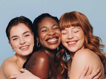 Gruppe glücklicher Frauen mit unterschiedlichen Hauttönen, die lächeln und sich in einem Studio umarmen | © Getty Images/jacoblund
