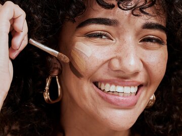 Eine junge Frau malt Make-up-Streifen mit einem Pinsel auf ihr Gesicht | © GettyImages/PeopleImages