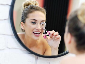 Frau sieht in den Spiegel und benutzt Wimpernzange | © Getty Images/macniak
