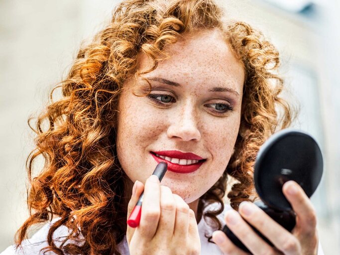 Rothaarige, junge Frau mit Locken blickt in ihren Handspiegel und trägt lächelnd Lippenstift auf | © Getty Images/Westend61