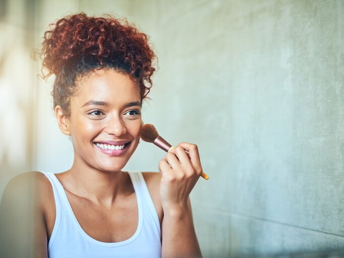 Junge Frau steht lächelnd vor dem Spiegel und trägt Makeup auf | © Getty Images/Moyo Studio