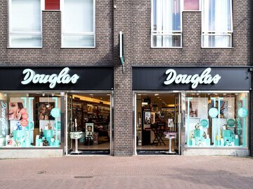 ALT-Text: "Die Vorderansicht eines Douglas-Kosmetikgeschäfts am Tag. Das Geschäft hat zwei große Schaufenster, die mit Werbung für Schönheitsprodukte und Angeboten dekoriert sind. In der Mitte befindet sich der Eingang, über dem in großen weißen Buchstaben 'Douglas' steht. | © Adobe Stock/Björn Wylezich