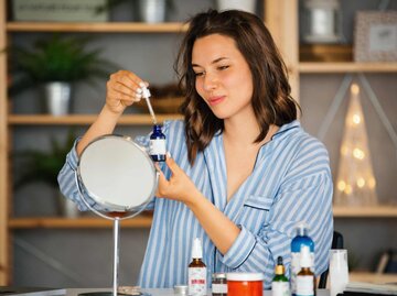 Frau mit Cremedosen und Serum in der Hand vor dem Spiegel | © Getty Images/Aja Koska