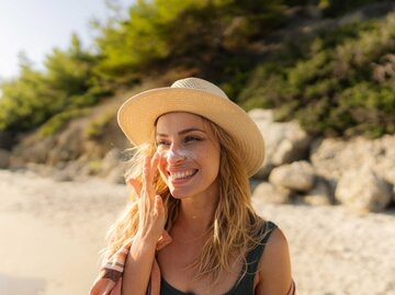 Frau mit Sonnenhut trägt Sonnencreme auf ihrer Nase auf und lächelt | © Getty Images/AleksandarNakic