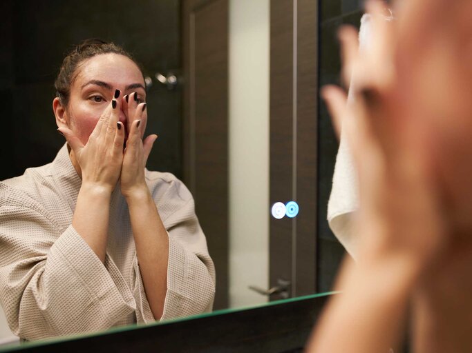 Frau steht im Bademantel vor dem Spiegel und massiert ihr Gesicht mit den Händen. | © Getty Images/Sergey Mironov
