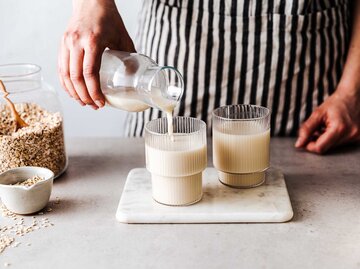 Frau mit Schürze schenkt am Küchentresen zwei Gläser Hafermilch ein. | © Getty Images / alvarez