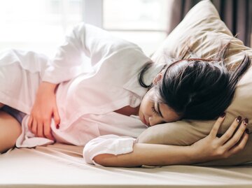 Frau liegt im Bett und fasst sich an den Unterleib vor Schmerzen | © Getty Images/Thnathip Pha Tir Wat'hn / EyeEm