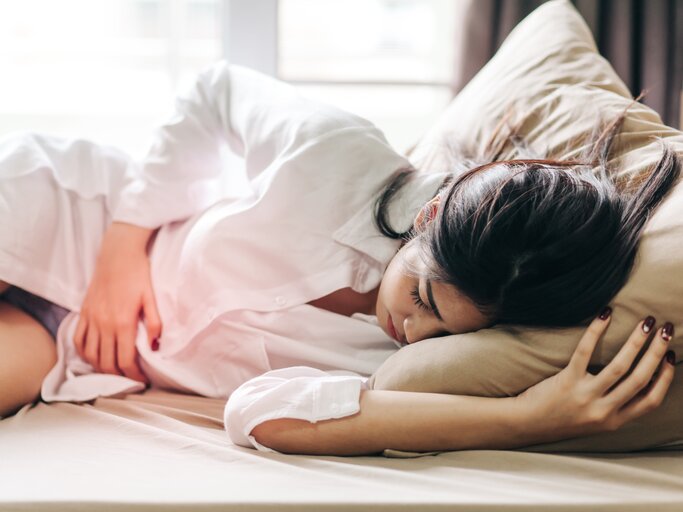 Frau liegt im Bett und fasst sich an den Unterleib vor Schmerzen | © Getty Images/Thnathip Pha Tir Wat'hn / EyeEm