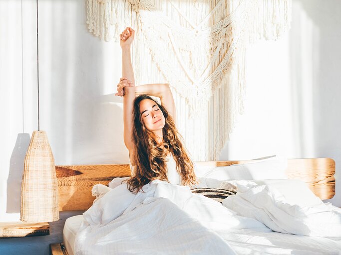 Eine junge Frau sitzt im Bett und streckt sich | © GettyImages/Maria Korneeva