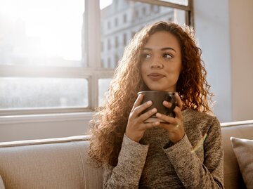 Eine junge Frau sitzt mit einer Kaffee Tasse in der Hand vor einem Fenster | © GettyImages/Delmaine Donson