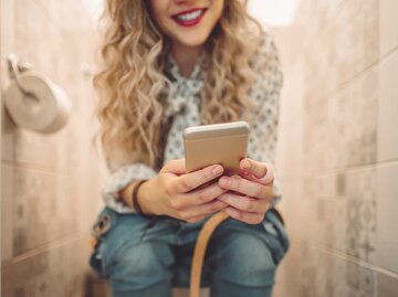 Frau sitzt auf der Toilette und schreibt auf ihrem Handy eine Nachricht. | © Getty Images / Eva-Katalin