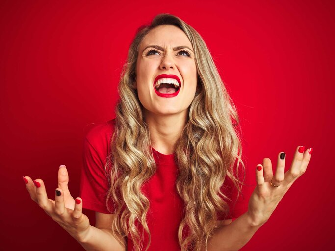 Wütende, blonde Frau in rotem T-Shirt schreit vor rotem Hintergrund | © Getty Images/AaronAmat
