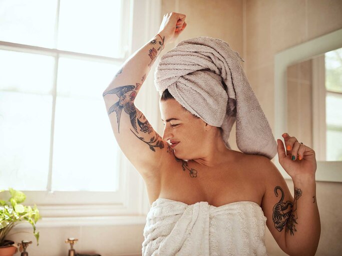 Frau steht mit Handtuch im Bad und riecht unter ihren Armen. | © Getty Images / PeopleImages