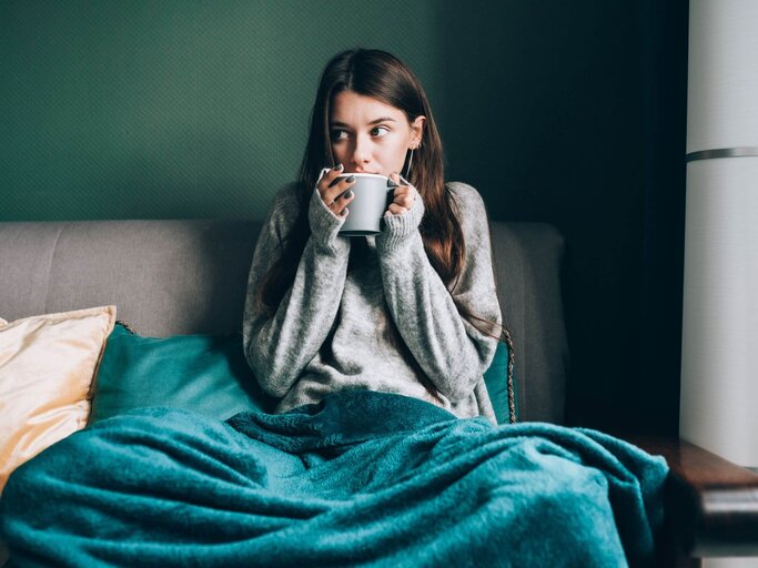 Person mit Teetasse und Decke auf Couch | © Getty Images/Olga Rolenko