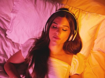Junge Frau liegt mit aufgesetzten Kopfhörern im Bett | © AdobeStock/Courtney/peopleimages.com