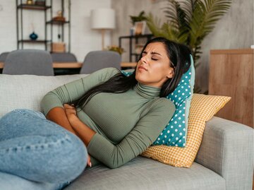 Frau liegt mit Unterleibsschmerzen auf der Couch | © Getty Images/urbazon