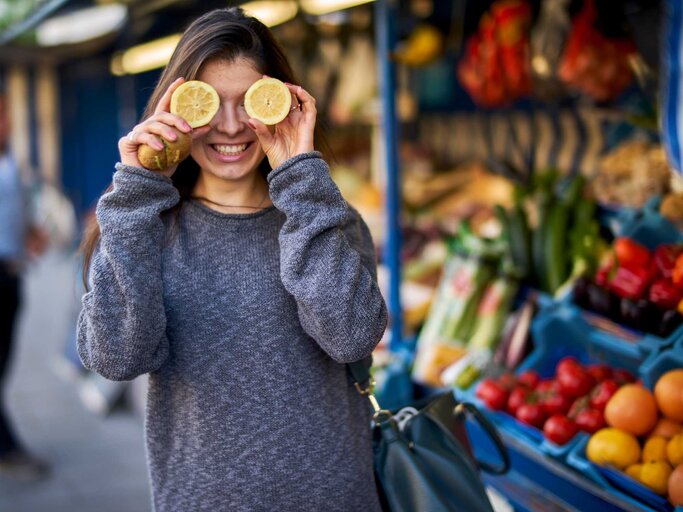 Frau auf einem Markt hält sich Zitronenhälften vor die Augen und lacht. | © Getty Images/ Westend61