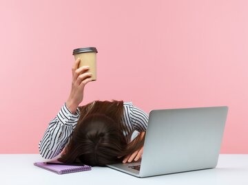 Arbeitende Frau liegt müde auf ihrem Laptop und hält einen Kaffee in der Hand | © gettyimages.de /Khosrork