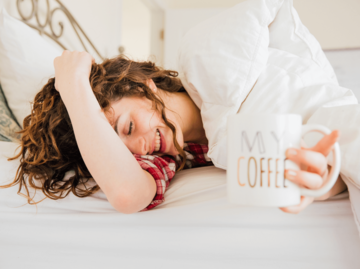 Frau liegt im Bett mit Kaffeetasse. | © Getty Images/Jena Ardell
