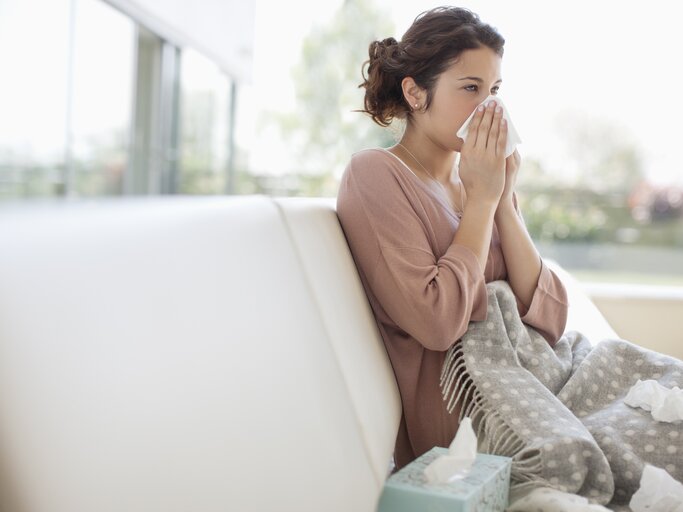 Frau sitzt erkältet auf der Couch und putzt sich die Nase | © Getty Images/Paul Bradbury