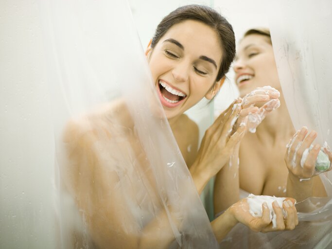 Zwei junge Frau lachen zusammen in der Badewanne hinter einem Duschvorhang | © Getty Images/PhotoAlto/Laurence Mouton