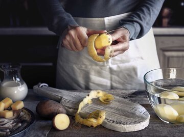 Frau steht mit Schürze in der Küche und schält Kartoffeln. | © Getty Images / KarpenkovDenis