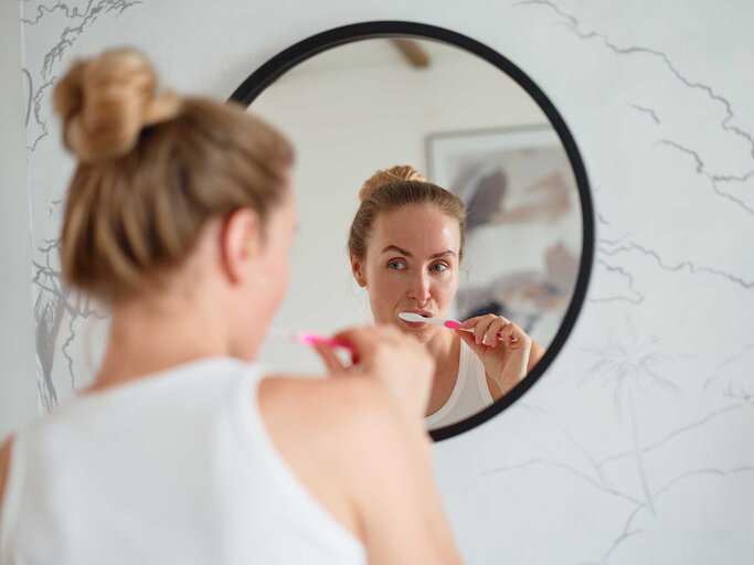 Frau mit Dutt steht vor einem runden Spiegel und putzt ihre Zähne. | © Getty Images / Sergey Mironov