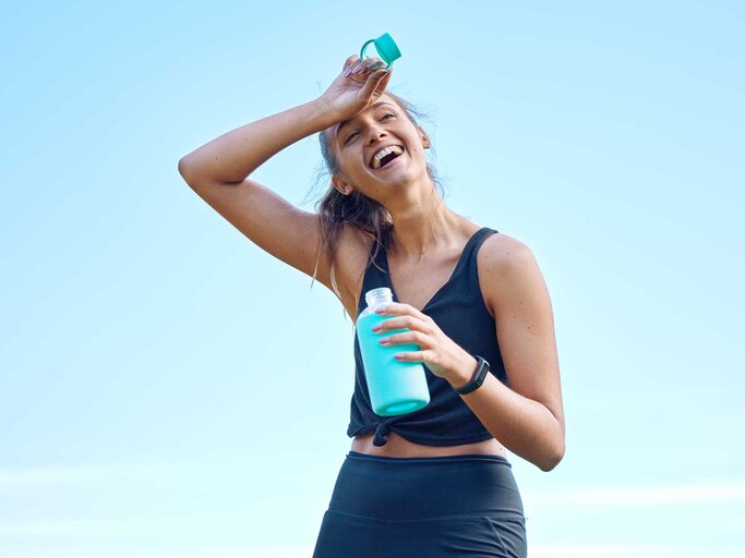 Junge Frau steht nach dem Sport mit einer Wasserflasche da und reibt sich den Kopf.  | © Getty Images / PeopleImages