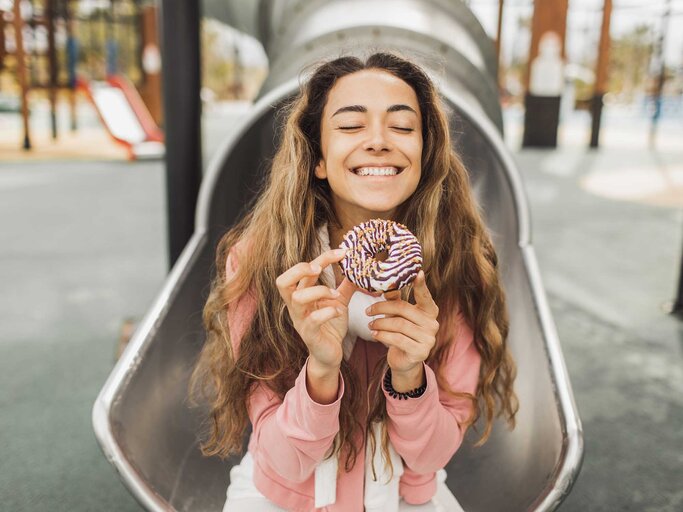 Junge Frau sitzt am Spielplatz mit einem Donut auf einer Rutsche. | © Getty Images / Oleg Breslavtsev