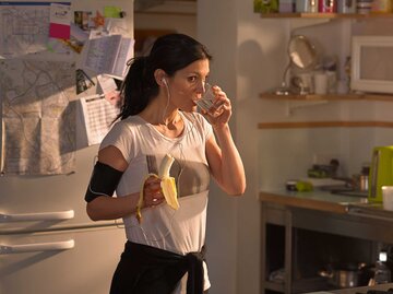 Frau steht morgens in der Küche und isst im Sportoutfit eine Banane und trinkt ein Glas Wasser. | © Getty Images / 10'000 Hours