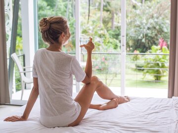 Frau sitzt am Morgen in ihrem Bett und trinkt ein Glas Wasser | © Getty Images/Jasmina007