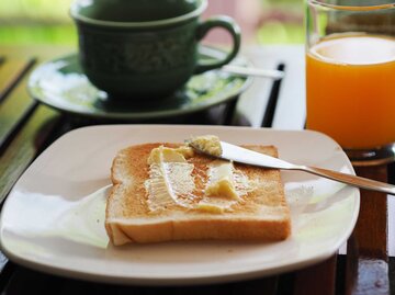 Auf einem Frühstückstisch mit Orangensaft und Kaffee liegt ein Toast mit Butter auf einem Teller. | © Getty Images / Penpak Ngamsathain