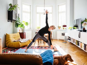 Frau steht in ihrem Wohnzimmer auf einer Yogamatte und macht die Yoga-Übung Dreieck. | © getty images / Tom Werner