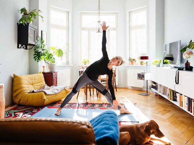 Frau steht in ihrem Wohnzimmer auf einer Yogamatte und macht die Yoga-Übung Dreieck. | © getty images / Tom Werner