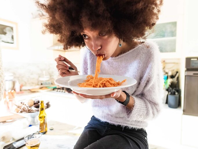 Junge Frau mit braunen Locken sitzt auf einer Küchenzeile und isst einen Teller Spaghetti mit Tomatensoße. | © Getty Images / Eugenio Marongiu