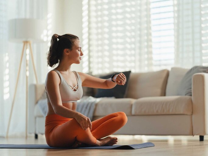 Junge Frau sitzt im Wohnzimmer auf der Yogamatte und schaut auf ihre Armbanduhr. | © Getty Images / Halfpoint Images