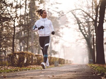 Junge Frau joggt im Winter mit einer weißen Jacke, schwarzen Leggings und Handschuhen durch eine Straße. | © Getty Images / martin-dm