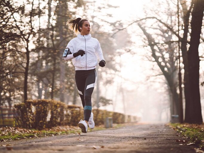 Junge Frau joggt im Winter mit einer weißen Jacke, schwarzen Leggings und Handschuhen durch eine Straße. | © Getty Images / martin-dm