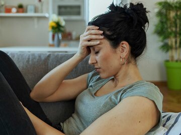 Frau sitzt erschöpft mit Kopfschmerzen auf der Couch | © Getty Images/PixelsEffect
