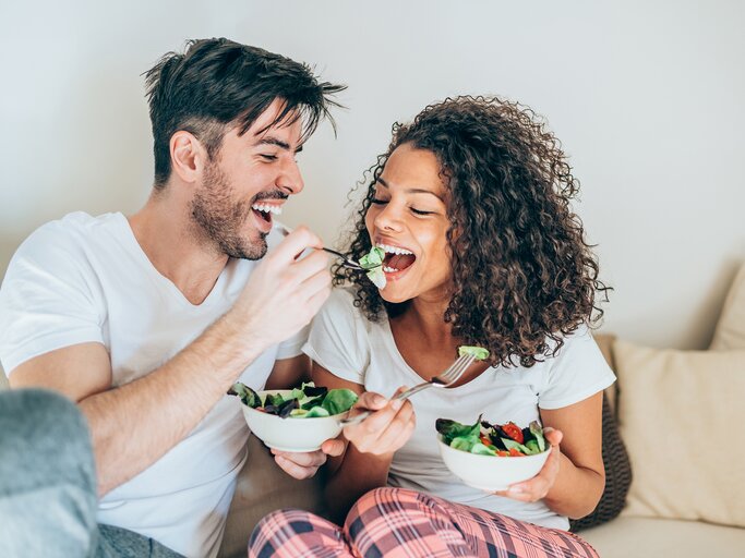 Ein Mann füttert eine Frau abends auf der Couch mit Salat | © GettyImages/VioletaStoimenova