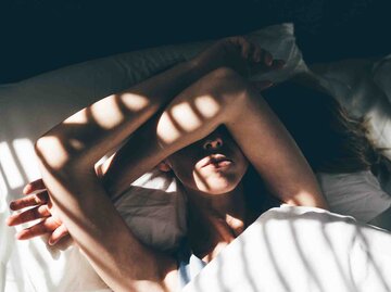 Frau liegt verschlafen im Bett und hält sich mit den Armen die Augen zu | © Getty Images/Maria Korneeva