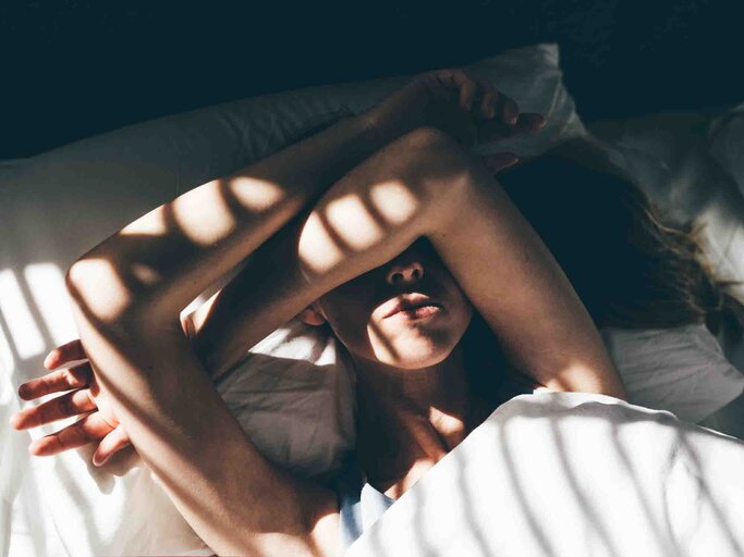 Frau liegt verschlafen im Bett und hält sich mit den Armen die Augen zu | © Getty Images/Maria Korneeva