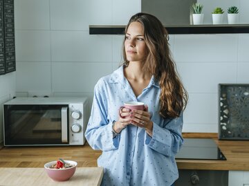 Frau steht in ihrer Küche mit einer Tasse in der Hand und schaut aus dem Fenster | © Getty Images/Westend61