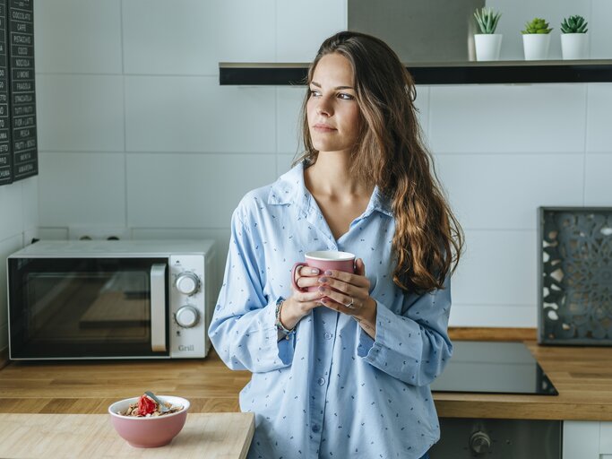 Frau steht in ihrer Küche mit einer Tasse in der Hand und schaut aus dem Fenster | © Getty Images/Westend61