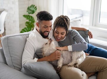 Junges Paar sitzt mit Hund auf der Couch und lächelt | © Getty Images/Halfpoint Images