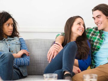 Frau sitzt mit ihrem Freund auf der Couch während ihre Freundin ihnen einen missbilligen Blick gibt | © Getty Images/JGI/Jamie Grill