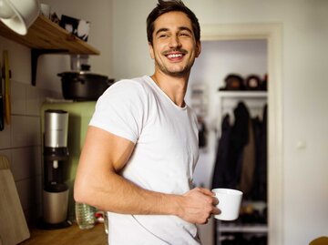 Junger Mann steht lächelnd mit einer Tasse in der Hand in seiner Küche | © Getty Images/Luis Alvarez