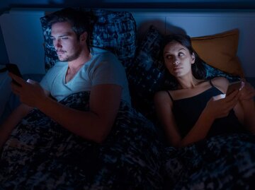 Frau liegt nachts im Bett mit ihrem Partner und schaut heimlich, was er sich an seinem Handy anschaut | © Getty Images/zoranm