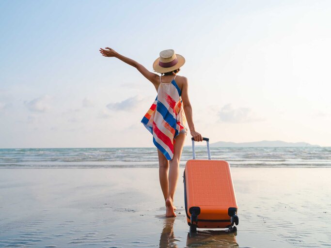 Frau steht mit ihrem Koffer am Strand und hebt euphorisch den Arm | © Getty Images/Twenty47studio