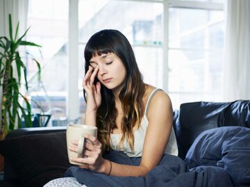 Müde wirkende Frau sitzt mit Kaffeetasse in der Hand auf dem Sofa | © Getty Images/Tara Moore